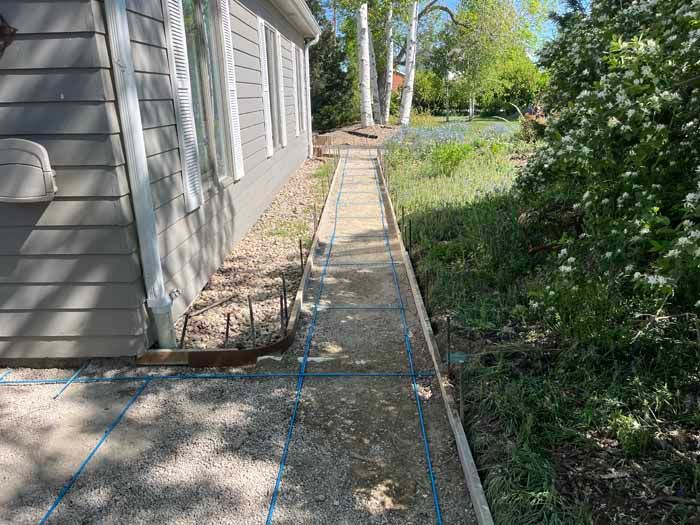 Sidewalk under construction next to a house and landscaping, with gravel and blue grid lines.
