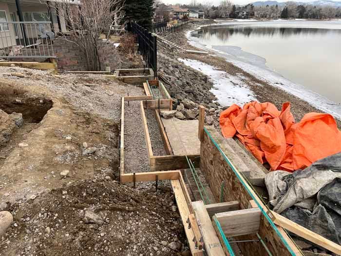 Construction site near a lake with wooden forms, rebar, gravel, and orange tarps.