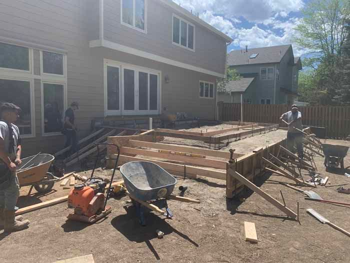 Construction workers building a concrete patio in a backyard. Forms, tools, and a home are visible.