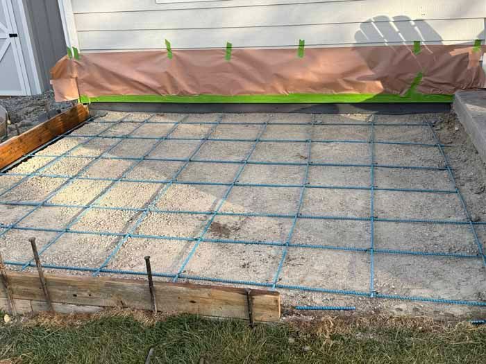 Concrete patio foundation with rebar grid laid on gravel base, near a house.