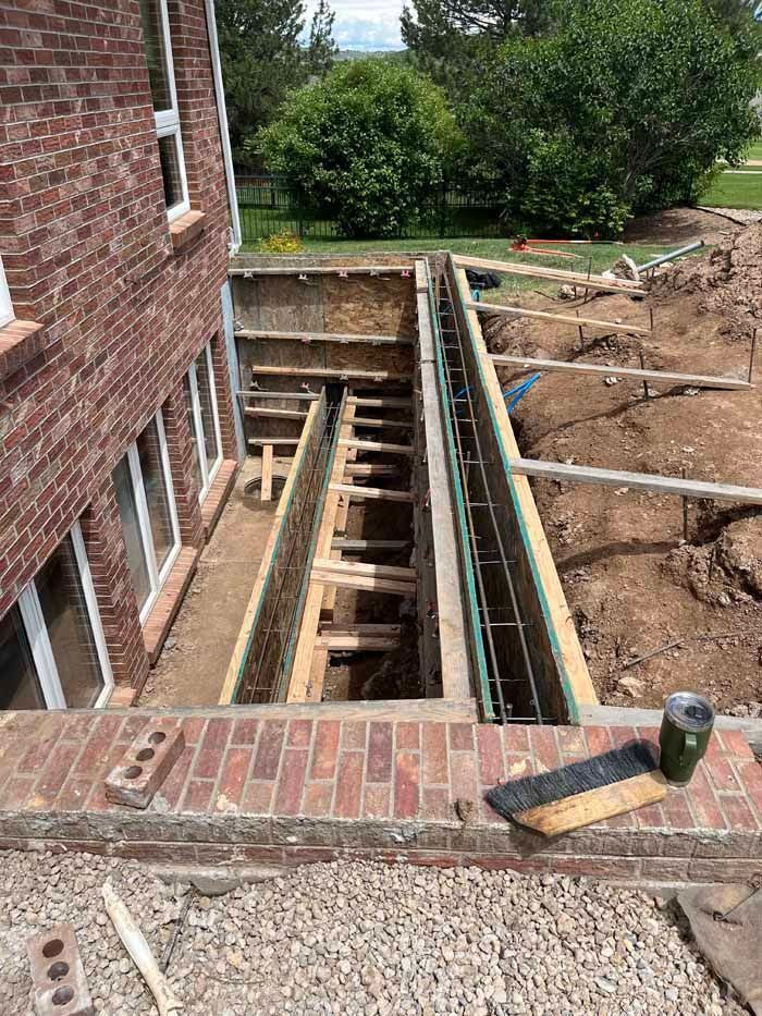 Construction site: basement window well. Excavation with wooden forms, brick wall, and building foundation visible.