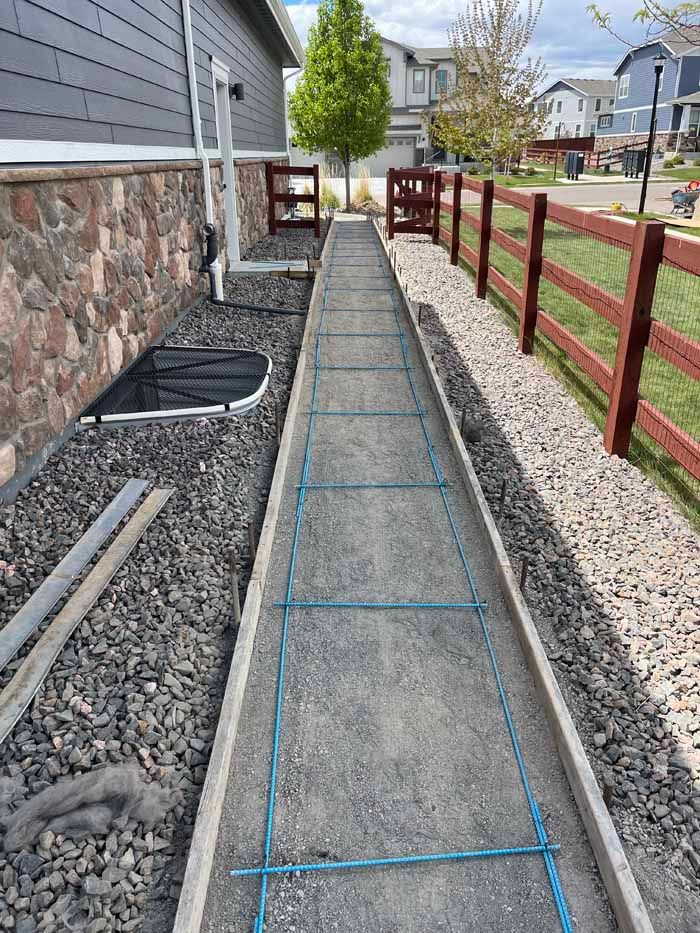 Concrete walkway under construction, bordered by gravel and a wooden fence. Rebar grid laid in the path.