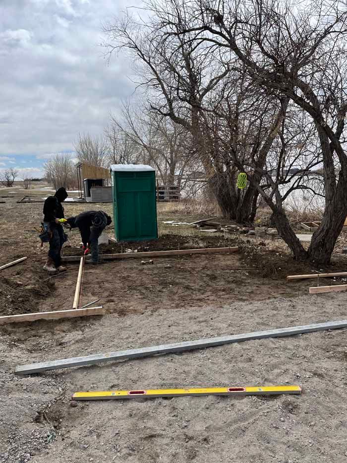 Two people building a wooden frame near a green outhouse and a leafless tree on a cloudy day.