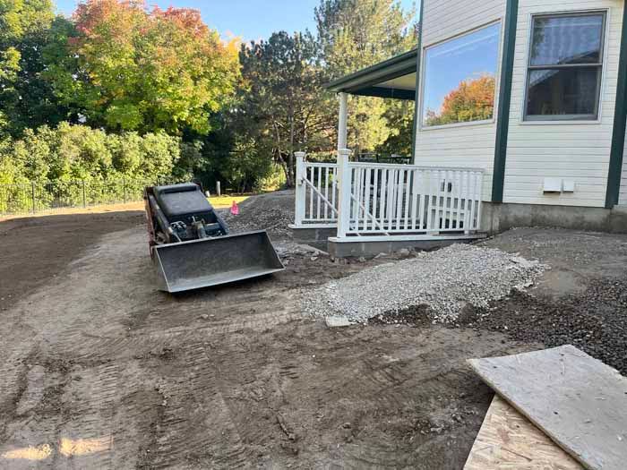 Mini excavator next to a house with a ramp and gravel pile, outdoor construction scene.