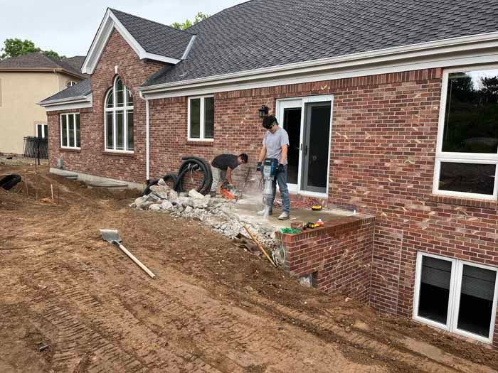Construction workers demolishing a brick patio attached to a red brick house with large windows.