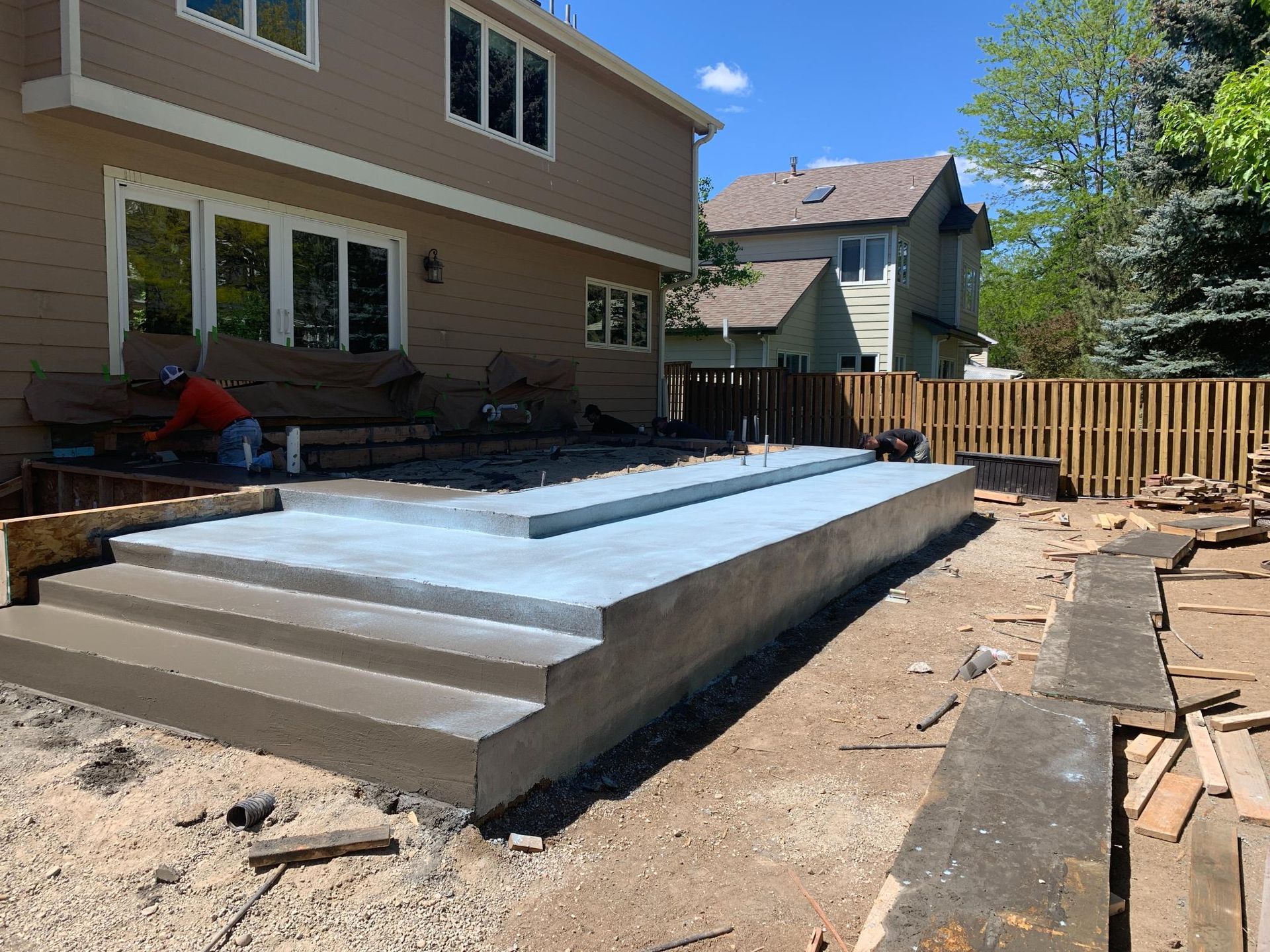 Concrete patio under construction with steps, next to a house and wooden fence.
