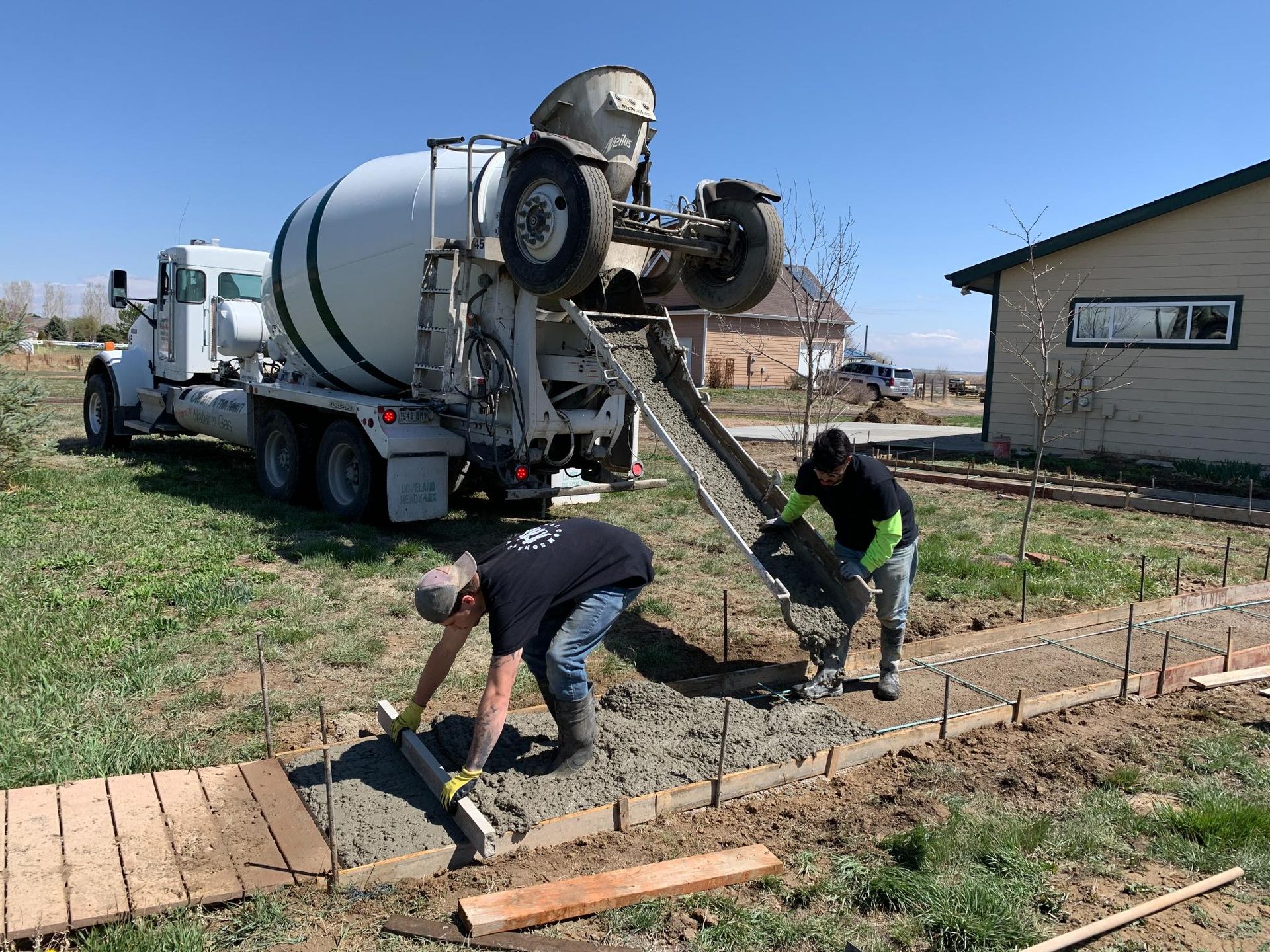 Concrete truck pouring concrete into a form for a sidewalk; two workers leveling the concrete.