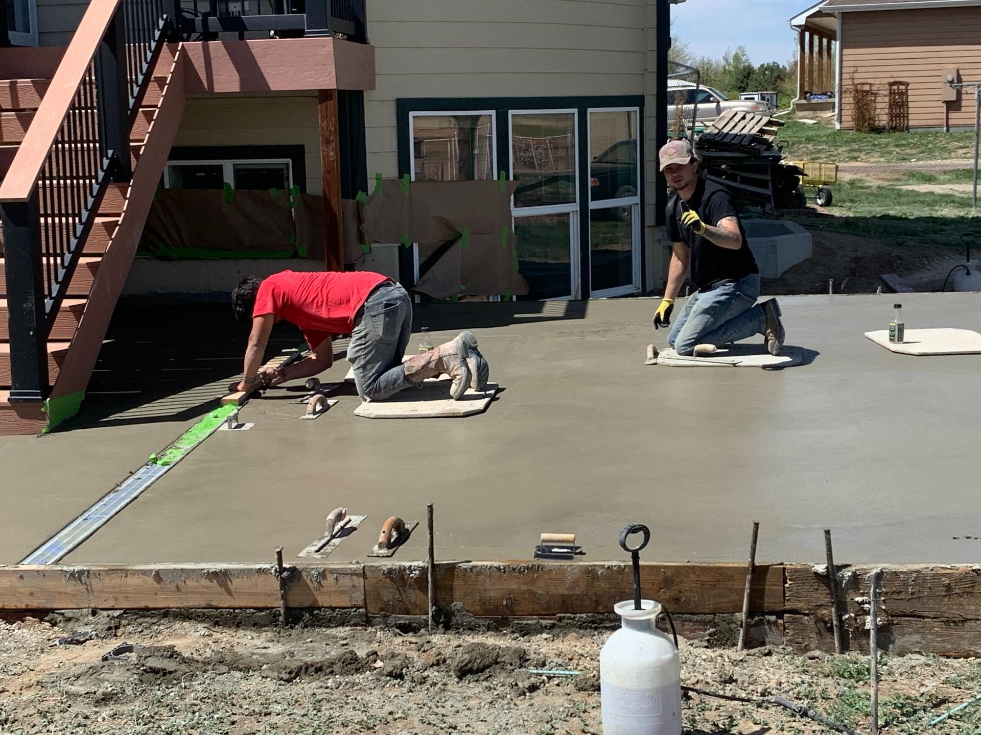 Two people smoothing wet concrete on a patio. Wooden frame, stairs, and a house in the background.