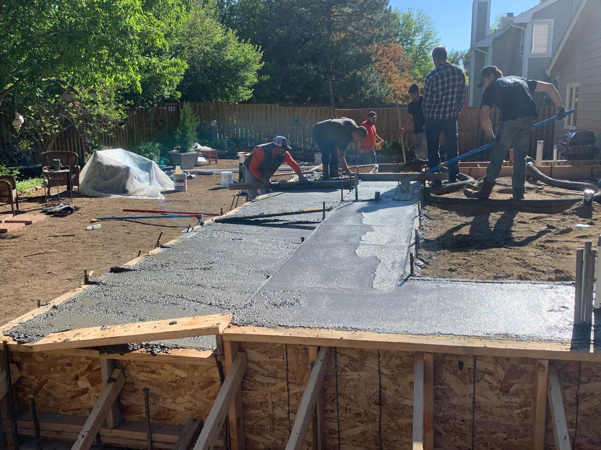 Construction workers pouring and smoothing concrete for a patio. Wooden forms, sunny outdoors, trees in background.