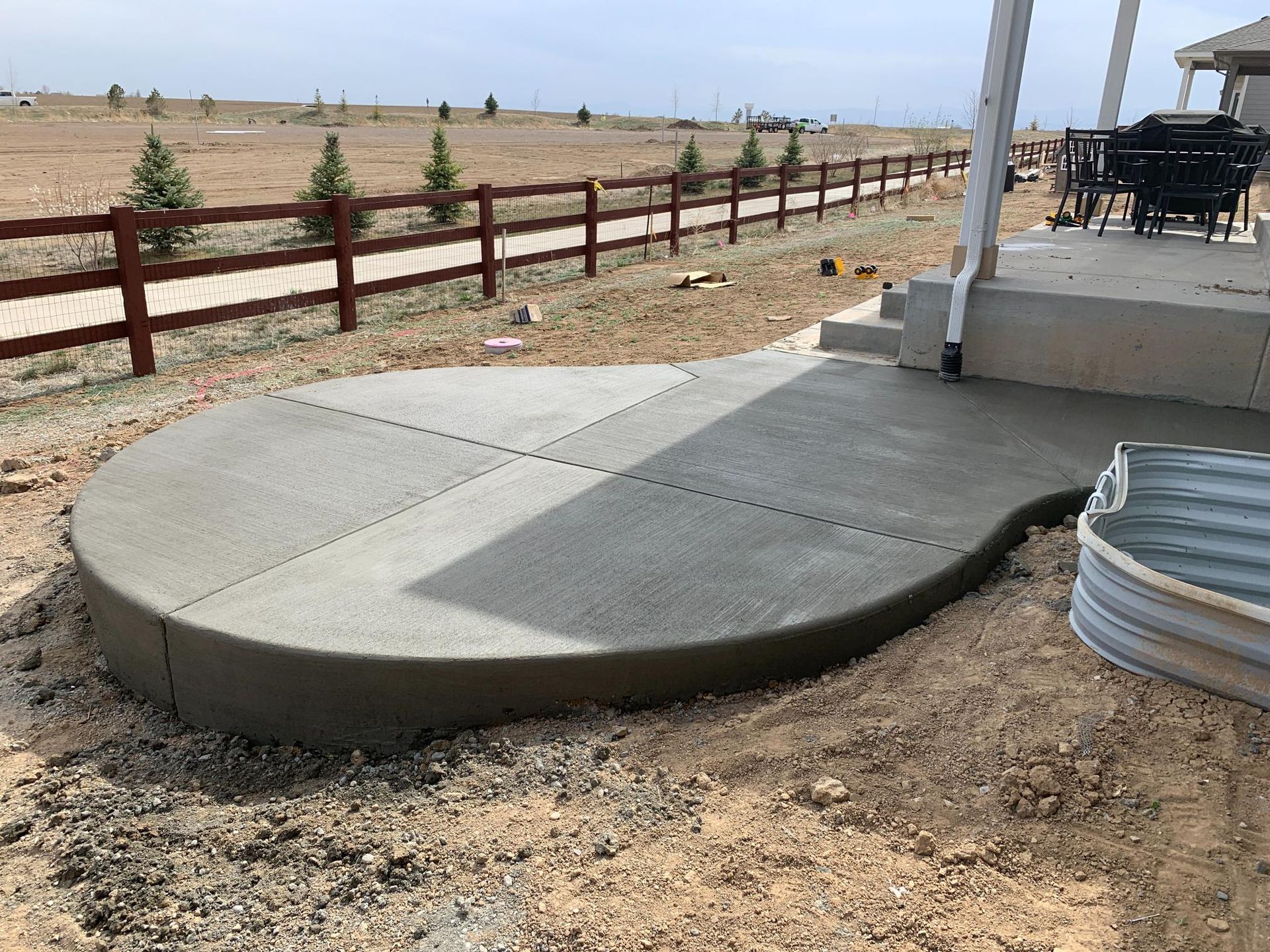 New concrete patio with rounded edge and scored sections, adjacent to covered porch and fence in a field.