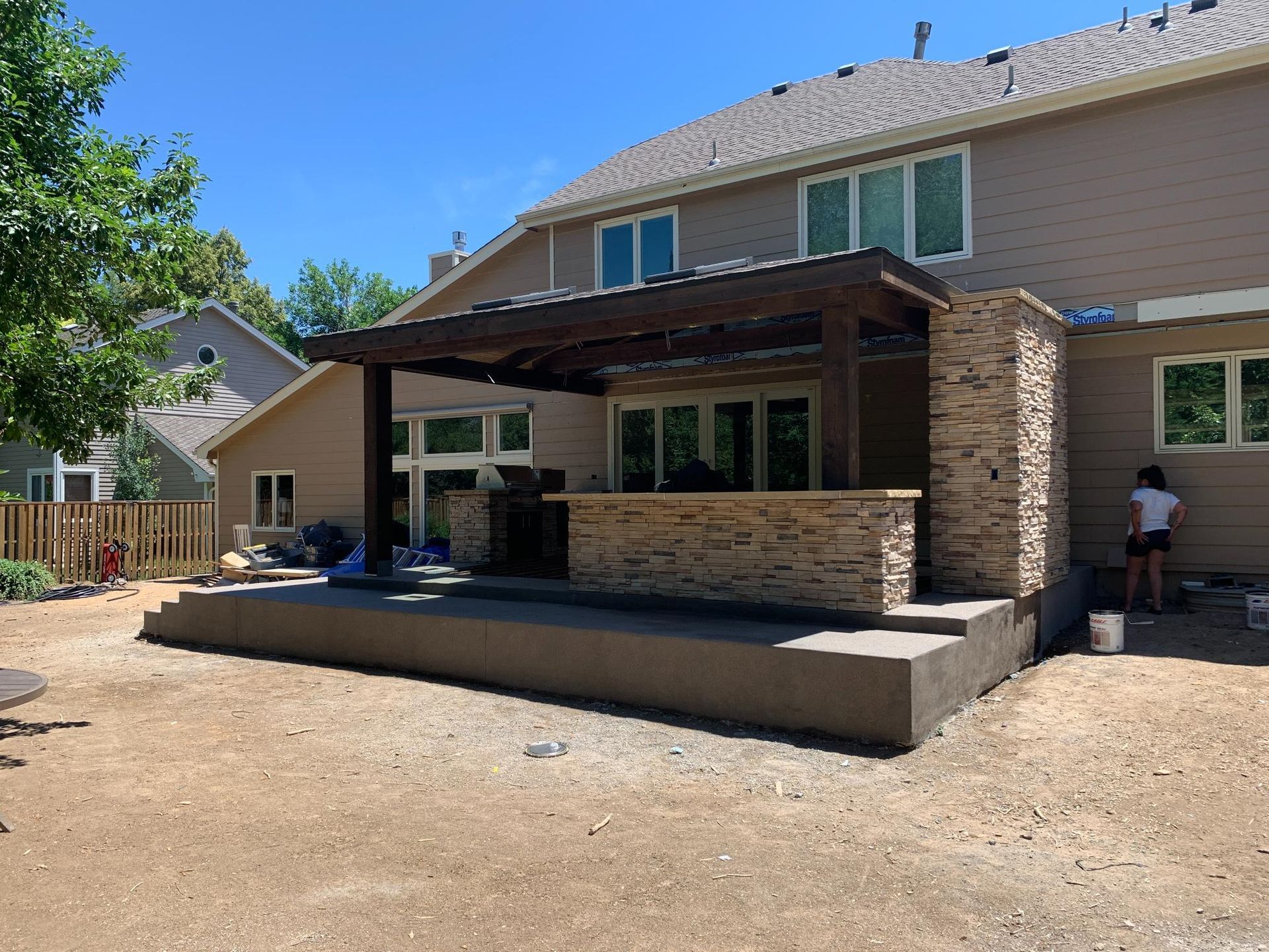 Backyard patio under construction with a pergola, stone accents, and a person working.