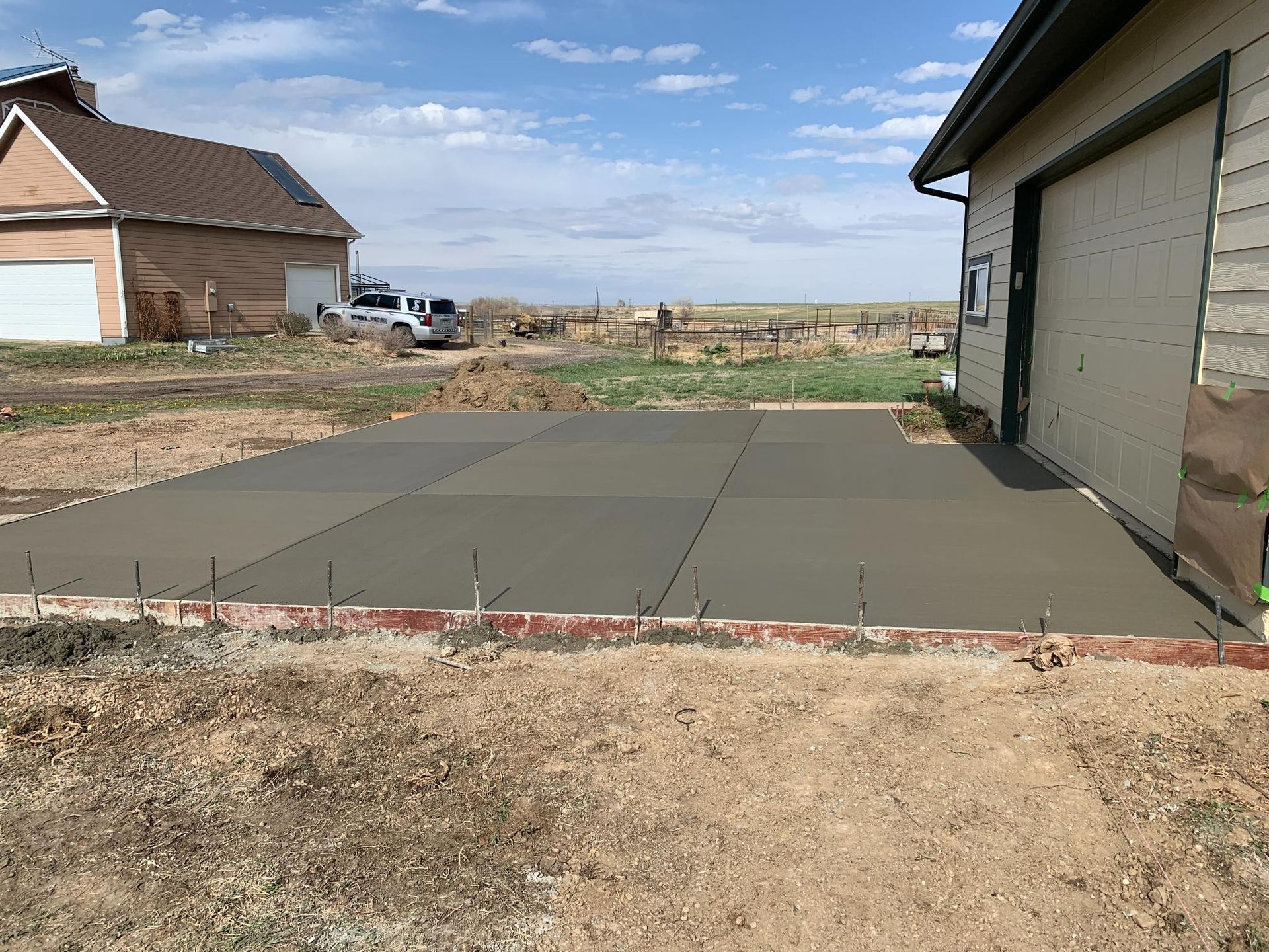 Newly poured concrete patio next to a tan garage and a red brick border, with a building in the background.