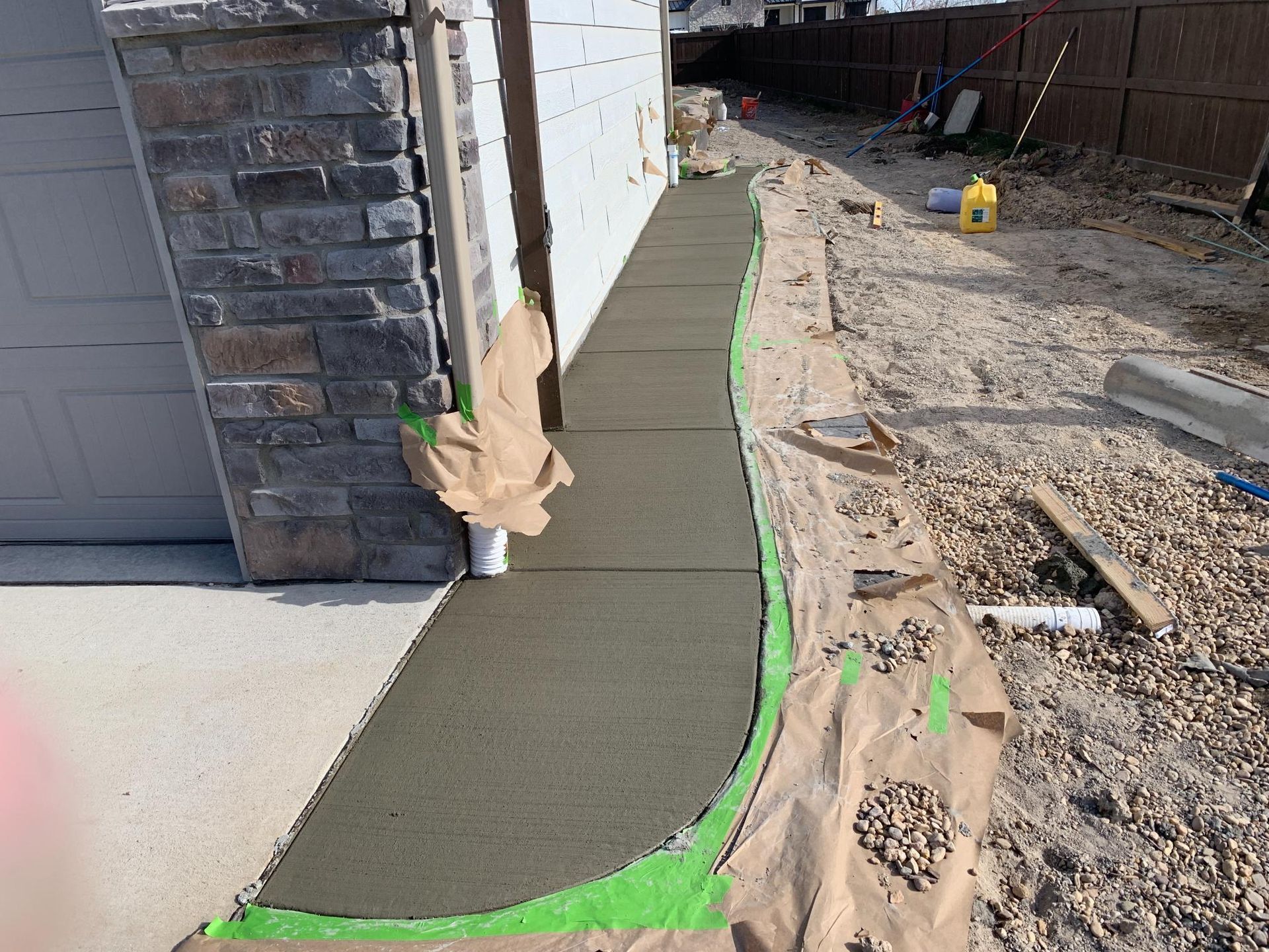 Freshly poured concrete sidewalk next to a building and gravel area. Paper and tape protect adjacent surfaces.