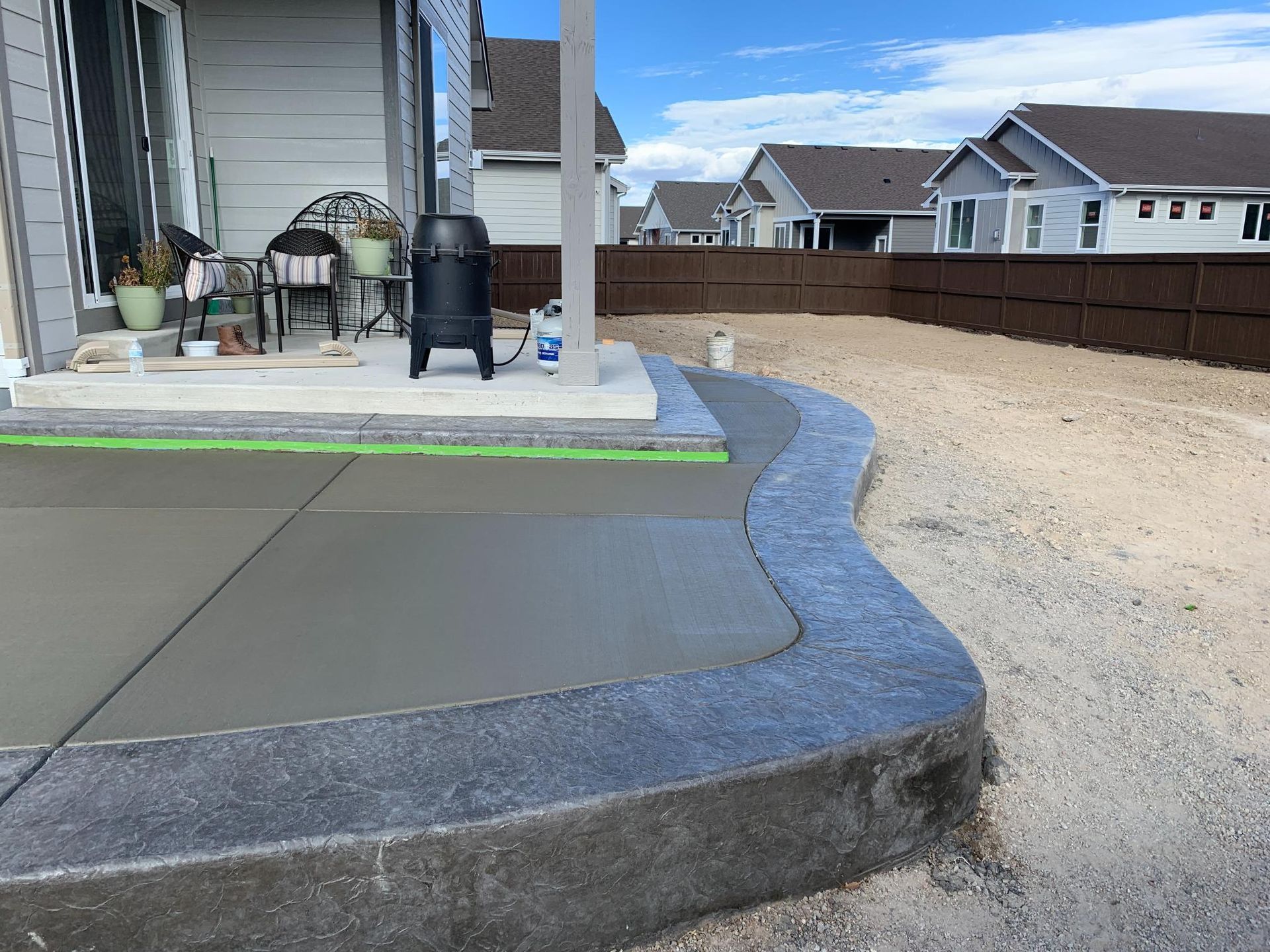 Newly poured concrete patio with a textured border, next to a house with gravel yard and brown fence.