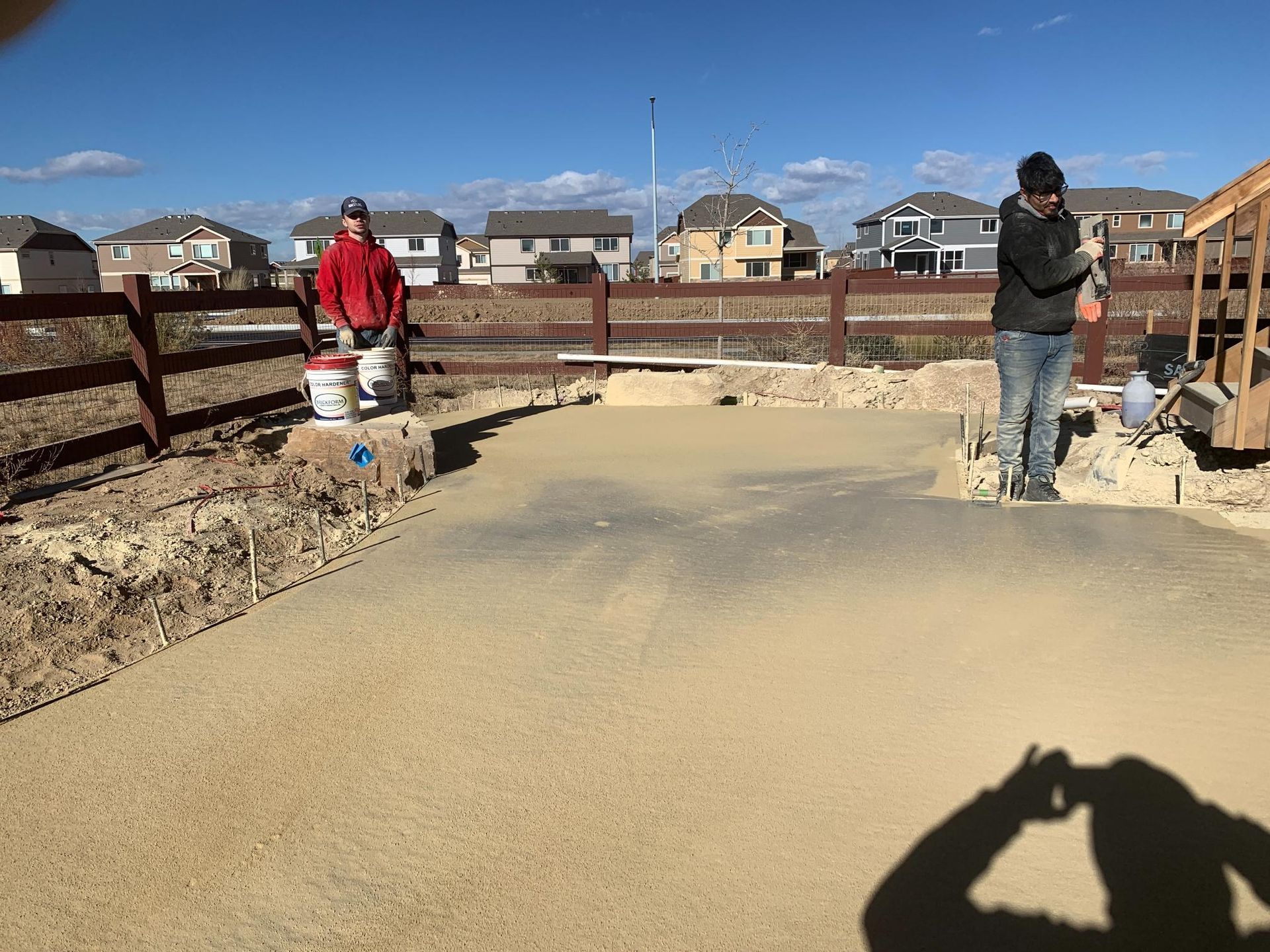 Two people working on a gravel surface next to a fence and houses.