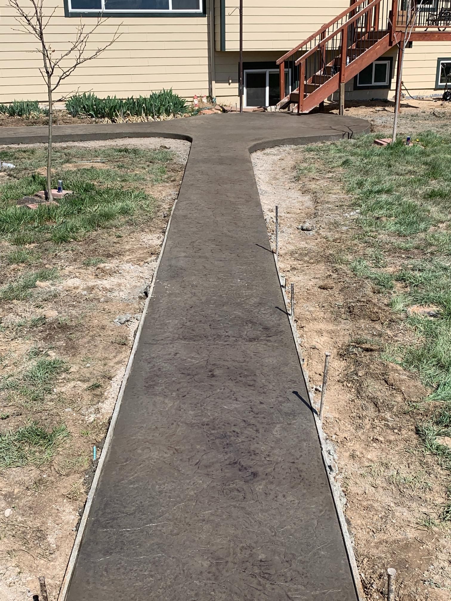 Newly poured concrete walkway leading to a house, flanked by exposed soil and stakes.