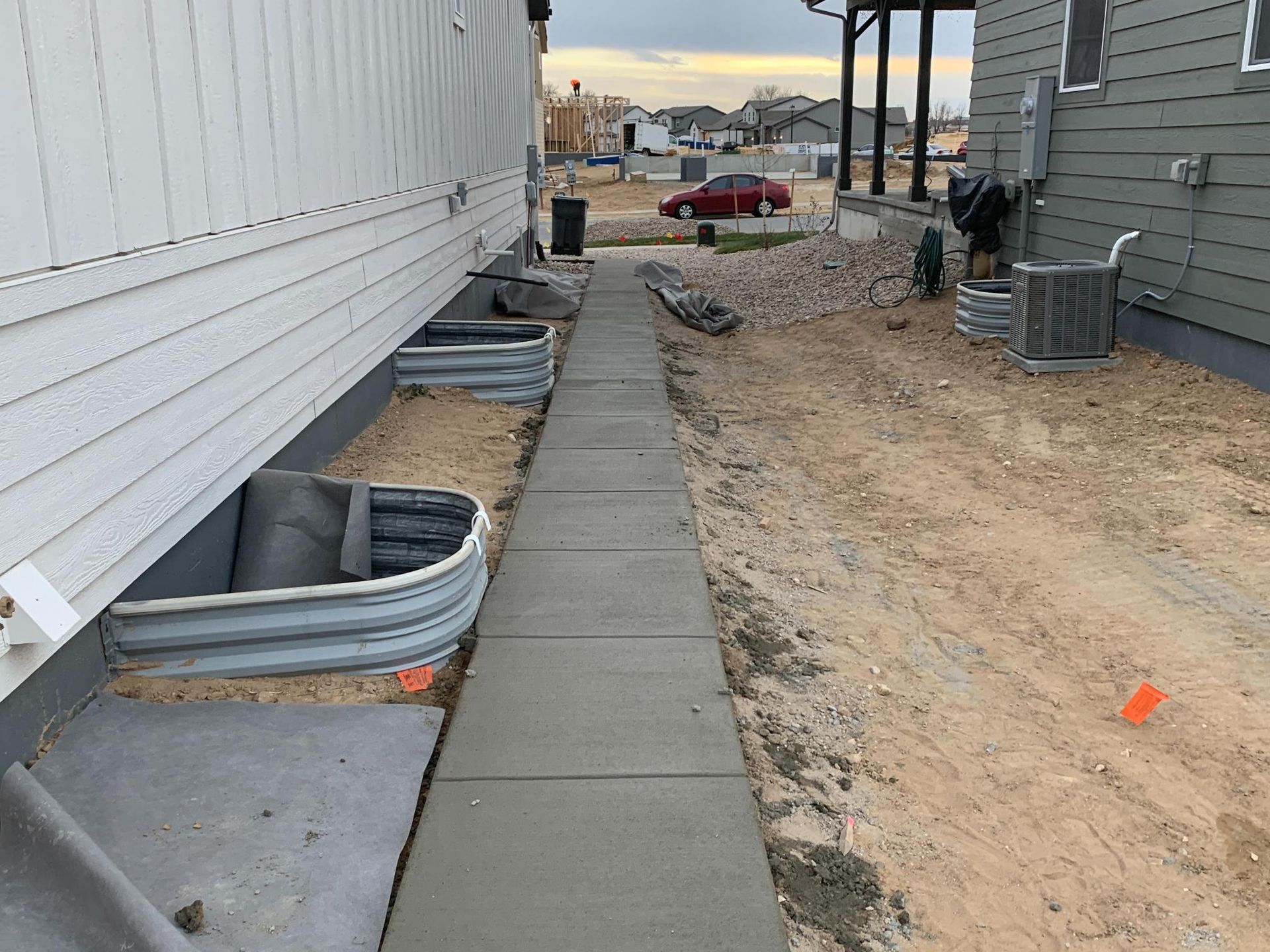 Cement walkway between two houses, one with window wells and the other with an AC unit, on a dirt lot.