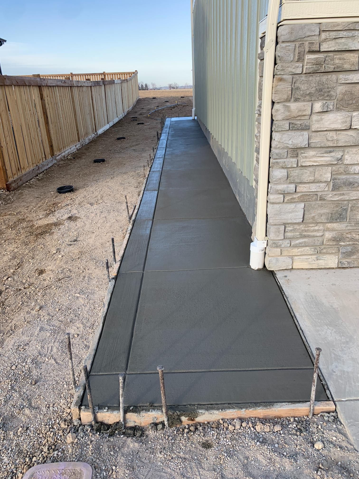 Newly poured concrete sidewalk next to a building and wooden fence. Construction materials visible.