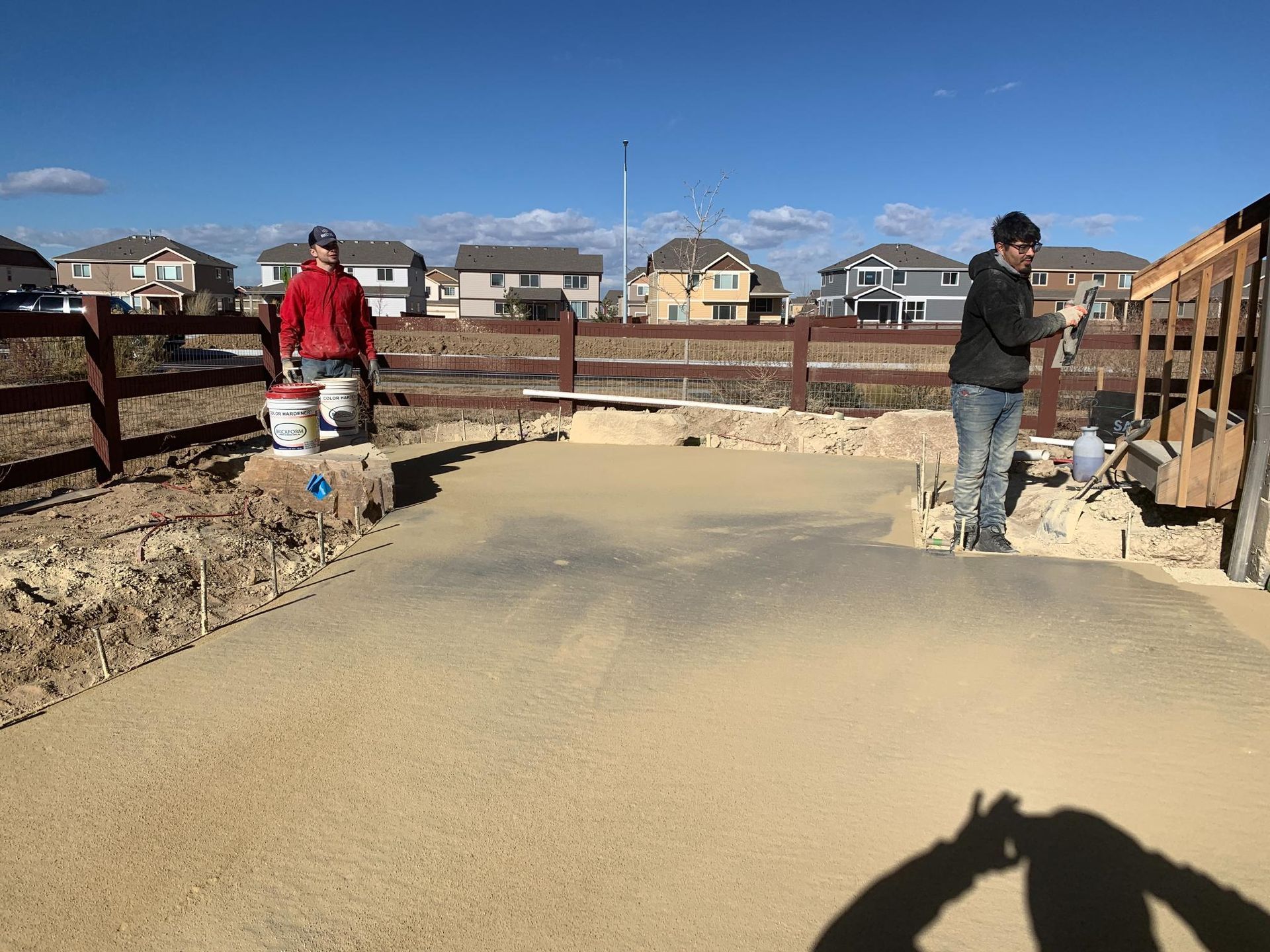 Two people painting a gravel area outdoors near a wooden fence, houses, and a clear blue sky.