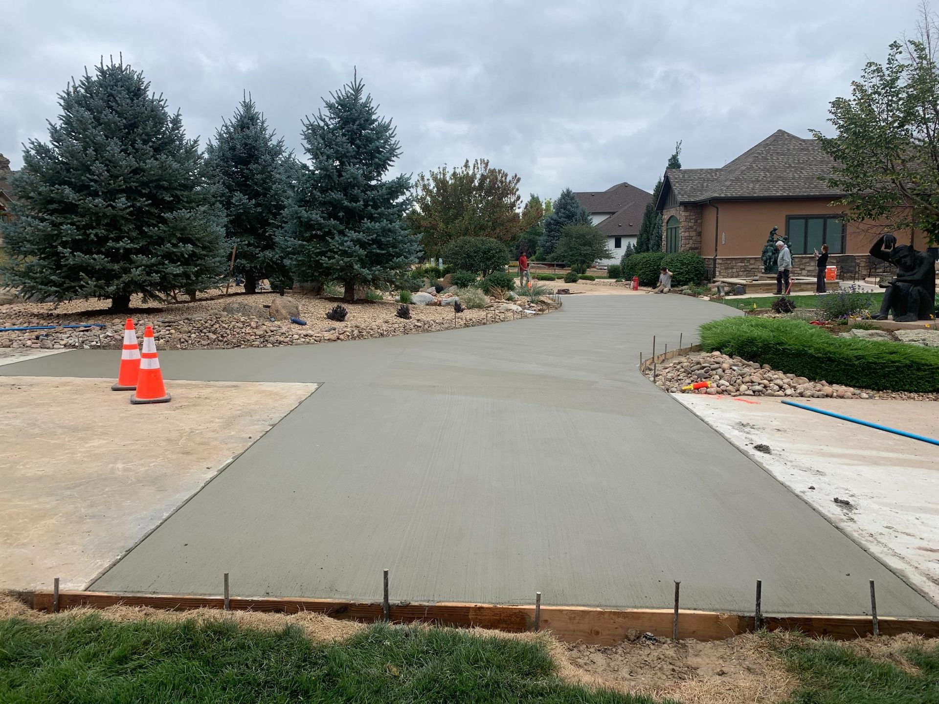 Newly poured concrete driveway with orange safety cones, trees, and house in background.