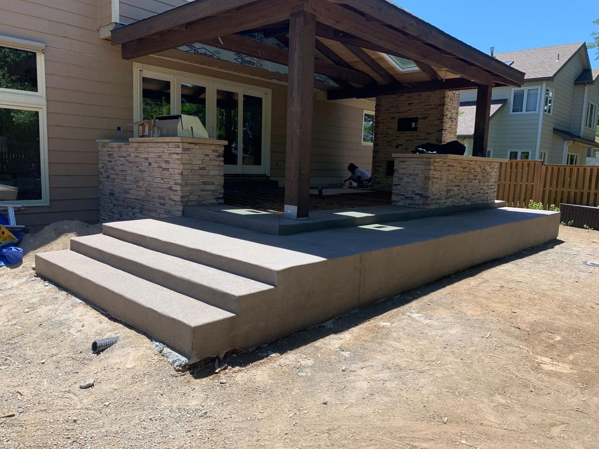 Concrete patio with steps, under a wooden pergola, adjacent to a house with large windows.