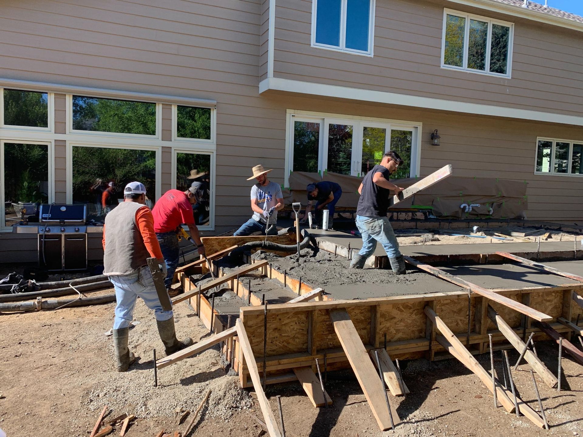 Construction workers pouring concrete for an outdoor structure next to a two-story beige house.