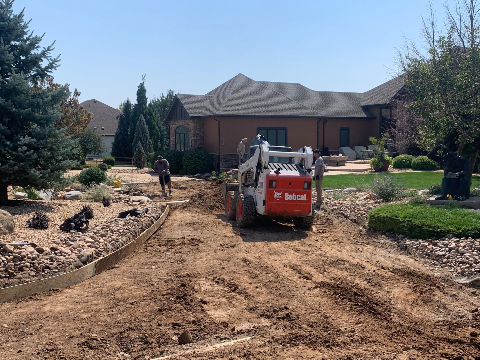 Bobcat compact loader on dirt road near a house, two workers, construction site, sunny.