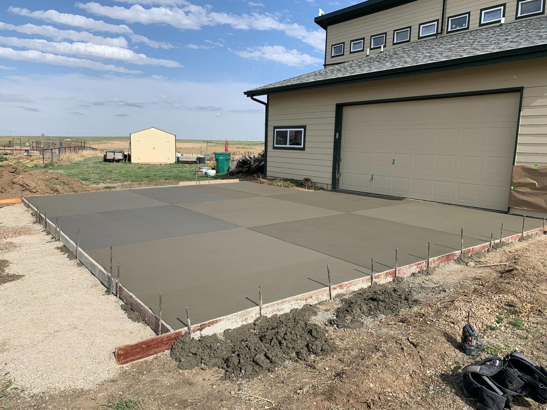 Newly poured concrete slab in front of a tan garage. Dirt and gravel surround the edges.