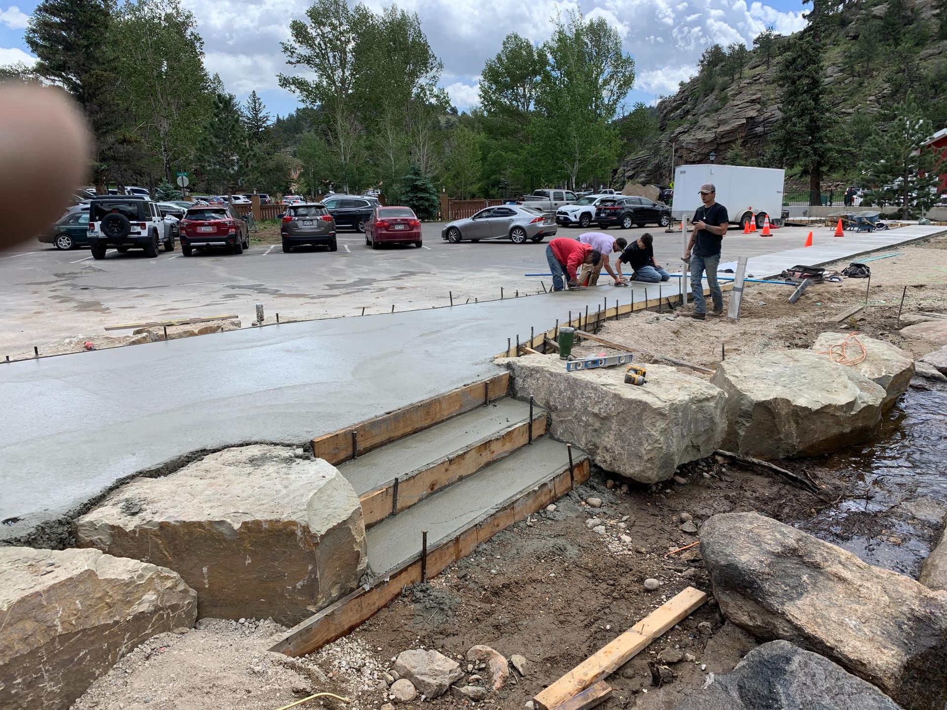 Concrete walkway with steps being built next to a stream. Workers and parked cars are visible.