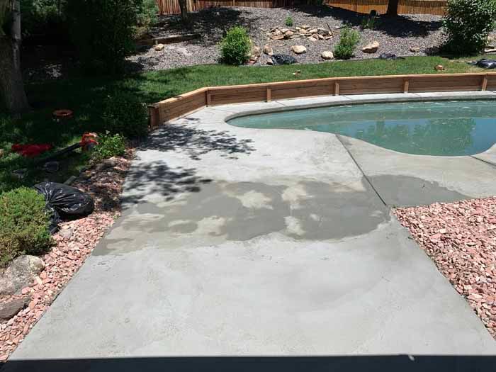 Concrete patio around a pool with wet, patched areas. Brown landscape border and red rock.