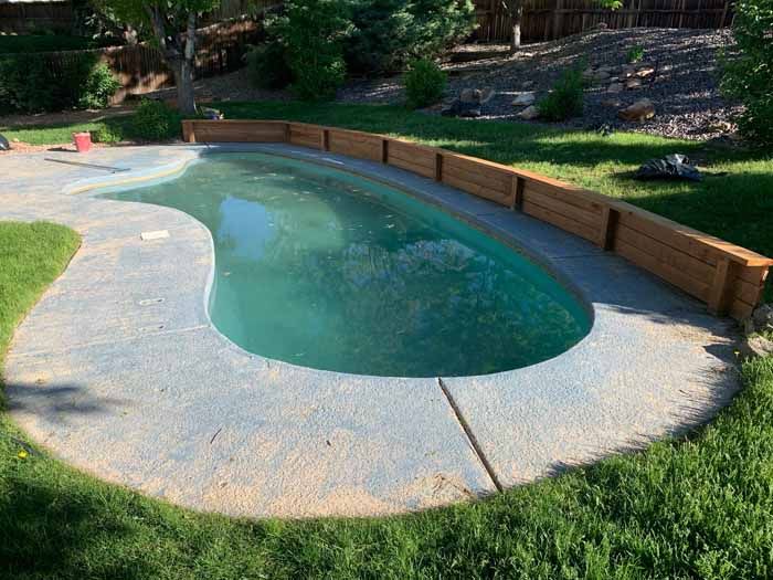 A pool with dirty water, surrounded by a concrete patio and a wooden retaining wall, on a grassy lawn.