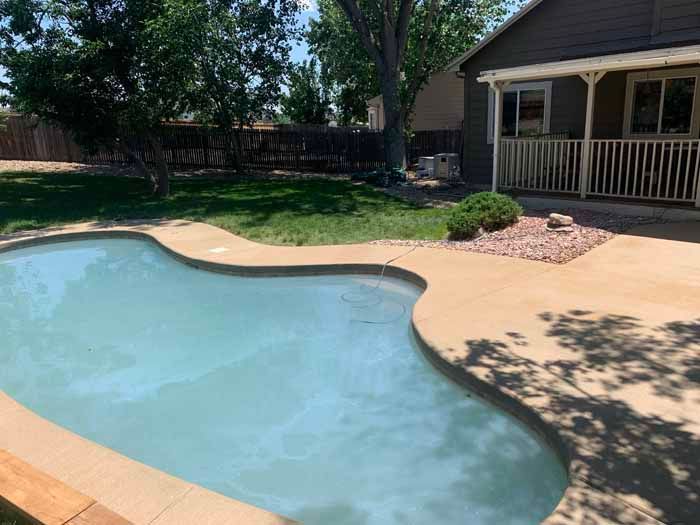 Backyard with light blue swimming pool, concrete patio, lawn, and house with porch.