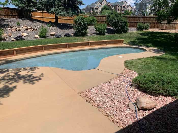 Swimming pool in a backyard with concrete patio, grass, wooden fence, and landscaping.
