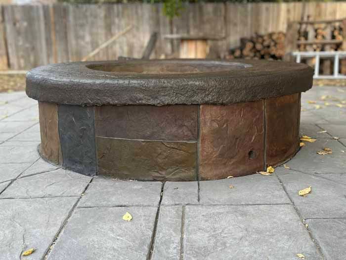 Stone fire pit on a patio, with a dark rim and multi-colored sides, surrounded by paving stones.