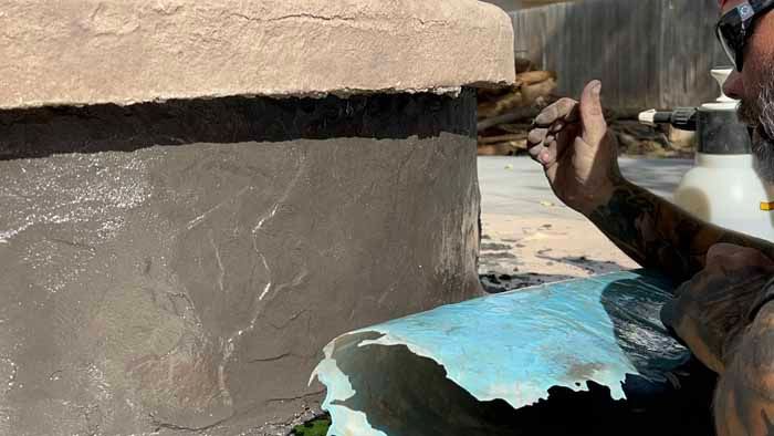 Person applying dark gray coating to a stone structure with a blue sheet in a sunny outdoor setting.