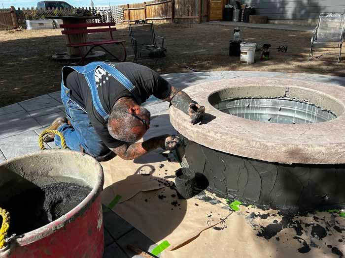 Man applying dark material to a circular structure, likely a fire pit. Kneeling outside, with materials nearby.
