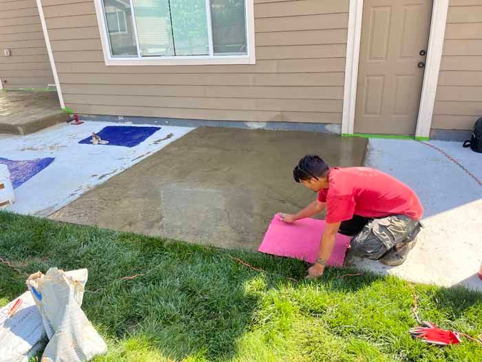 Person kneeling, placing a pink mat on a wet concrete patio near a house, bags of material nearby.