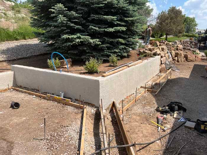 Construction of a concrete retaining wall with a large evergreen tree in the background, surrounded by dirt and landscape materials.