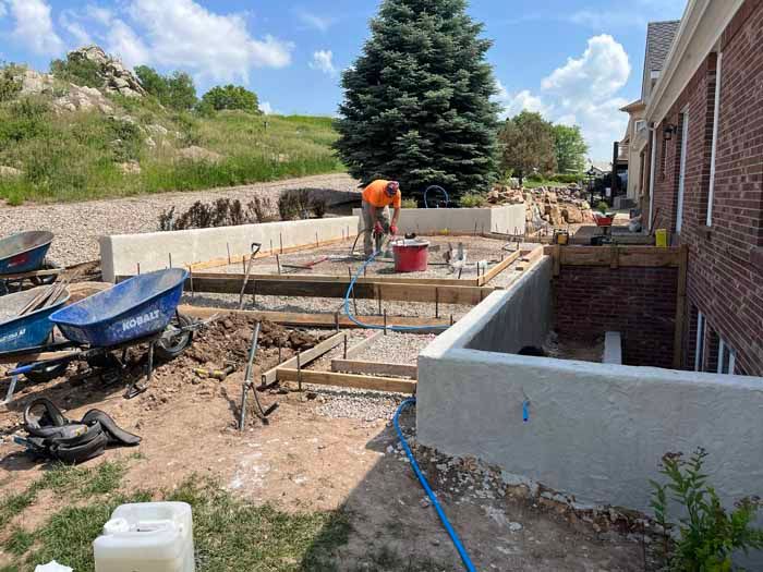 Construction site with worker using tools. Cement foundation, wheelbarrows, and retaining walls are visible.
