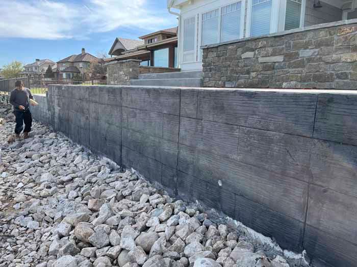 Stone retaining wall with a person nearby on a rocky shoreline, house in the background.