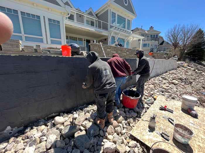 Construction workers applying stucco to a retaining wall near a lake, under a large house.