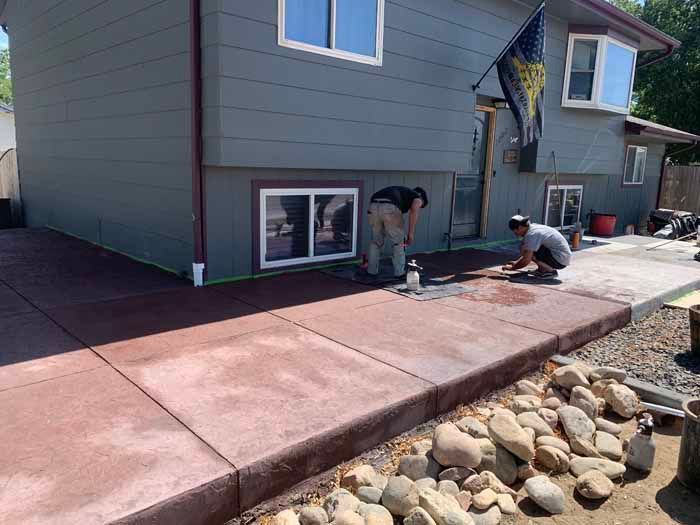 Workers finishing a stamped and stained concrete sidewalk next to a house with grey siding.