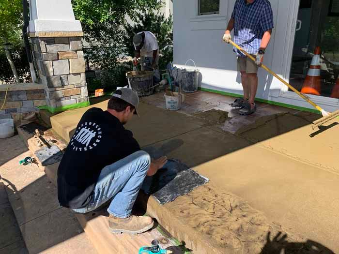 Workers applying concrete to a porch. One kneels, troweling; another spreads it with a squeegee; a third works on a stone column.