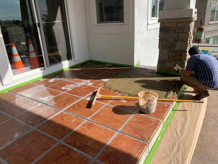 Man troweling cement on a porch floor. Red tiles, green tape, and a bucket are visible.