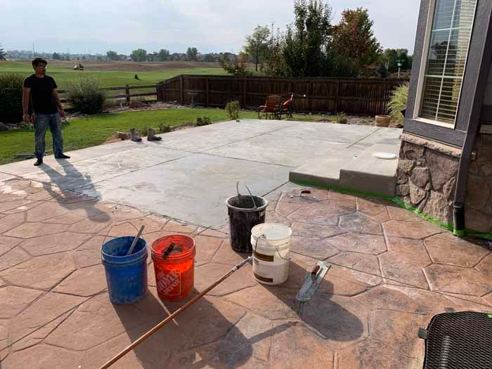 Man standing on a concrete patio being worked on. Buckets of paint and tools are scattered around. Green grass and a fence are in the background.