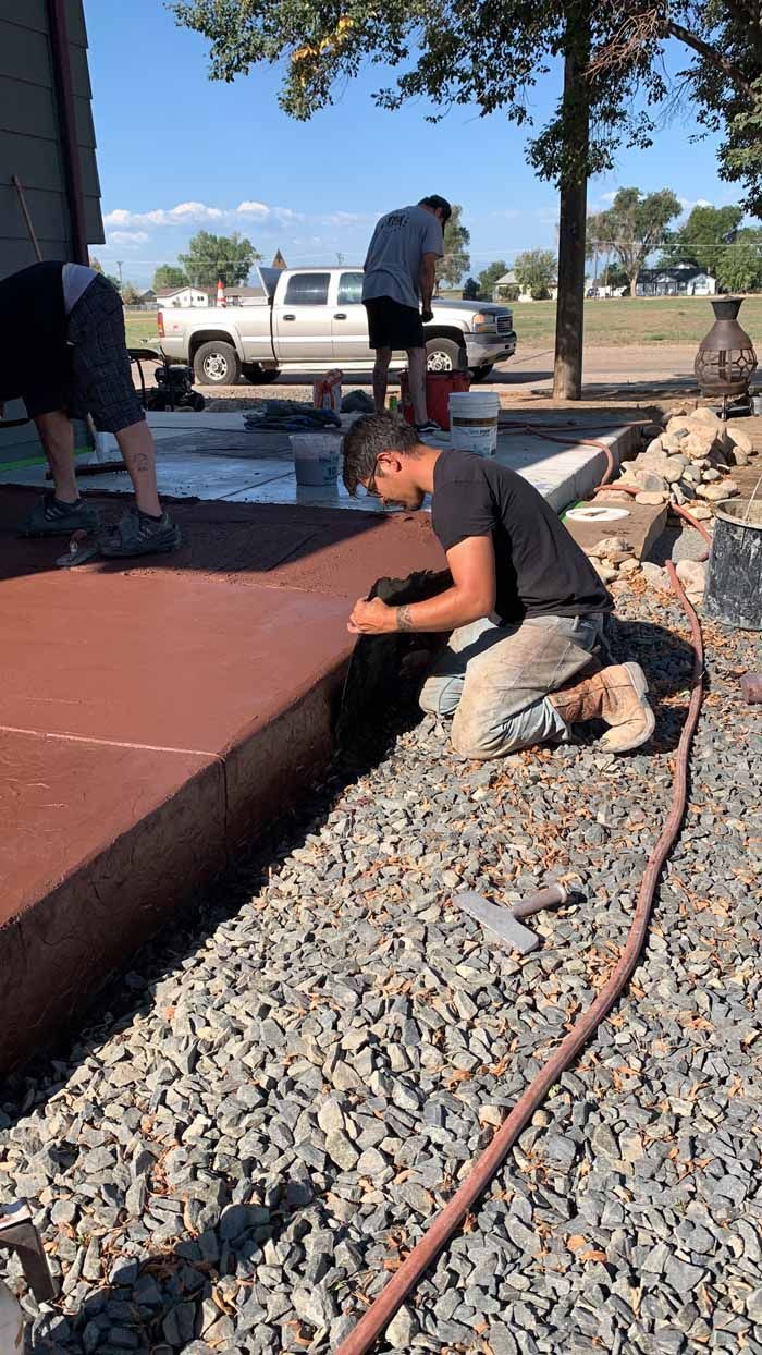 Three people working on a concrete project outdoors. One kneels, focused; two others stand nearby.
