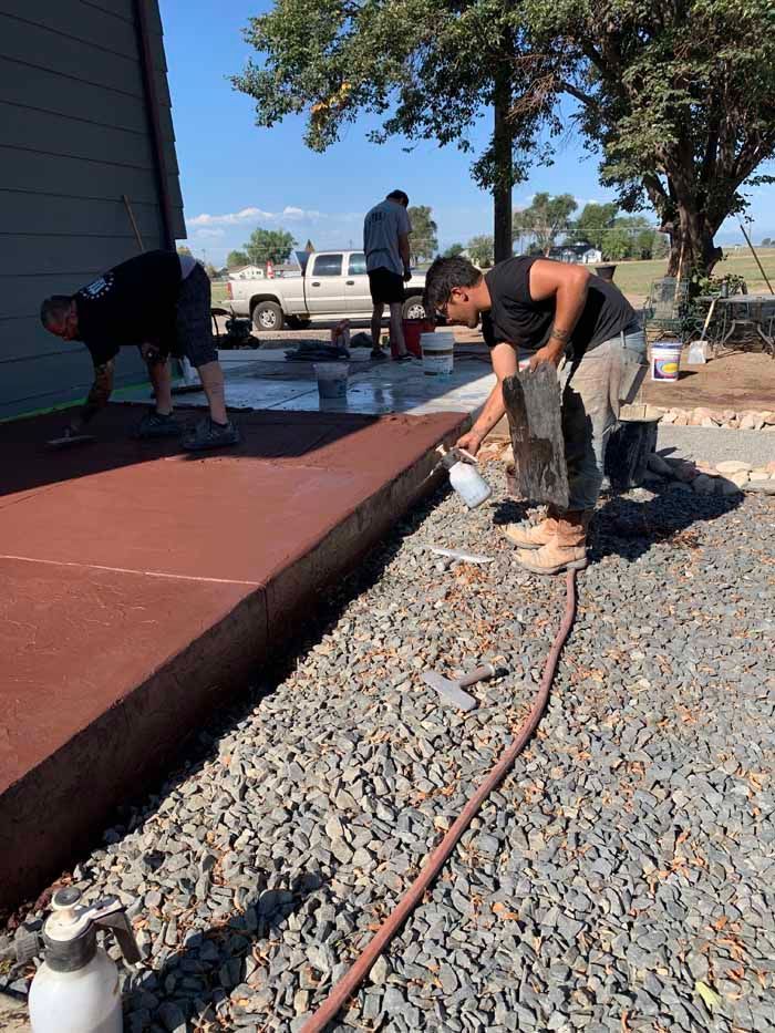Three people working on a concrete project outdoors. Two men are applying a reddish substance to the concrete, while another works in the background.