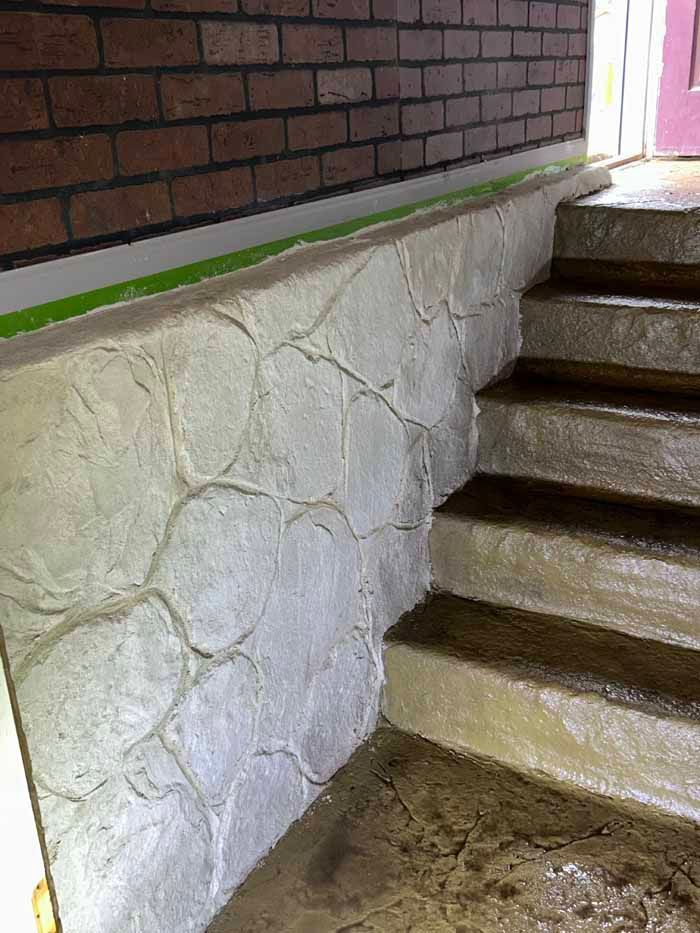 Stone-textured concrete wall beside damp concrete steps leading up to an entrance. Brick wall and green trim are also present.
