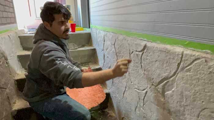Man applies finish to stone-like wall in a stairwell. He kneels and uses a tool.