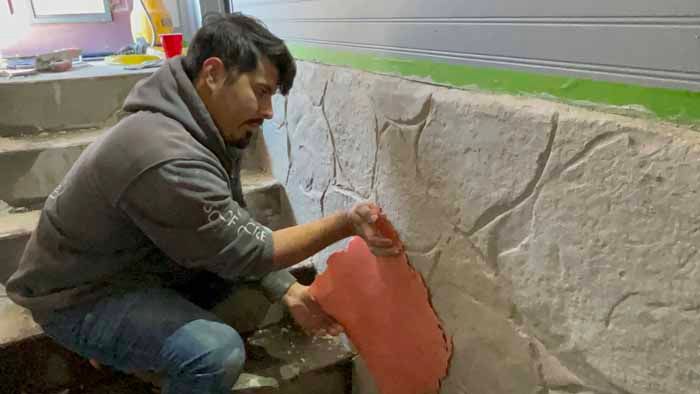 Man applying reddish material to stone wall. Outdoors with a gray hoodie and jeans.